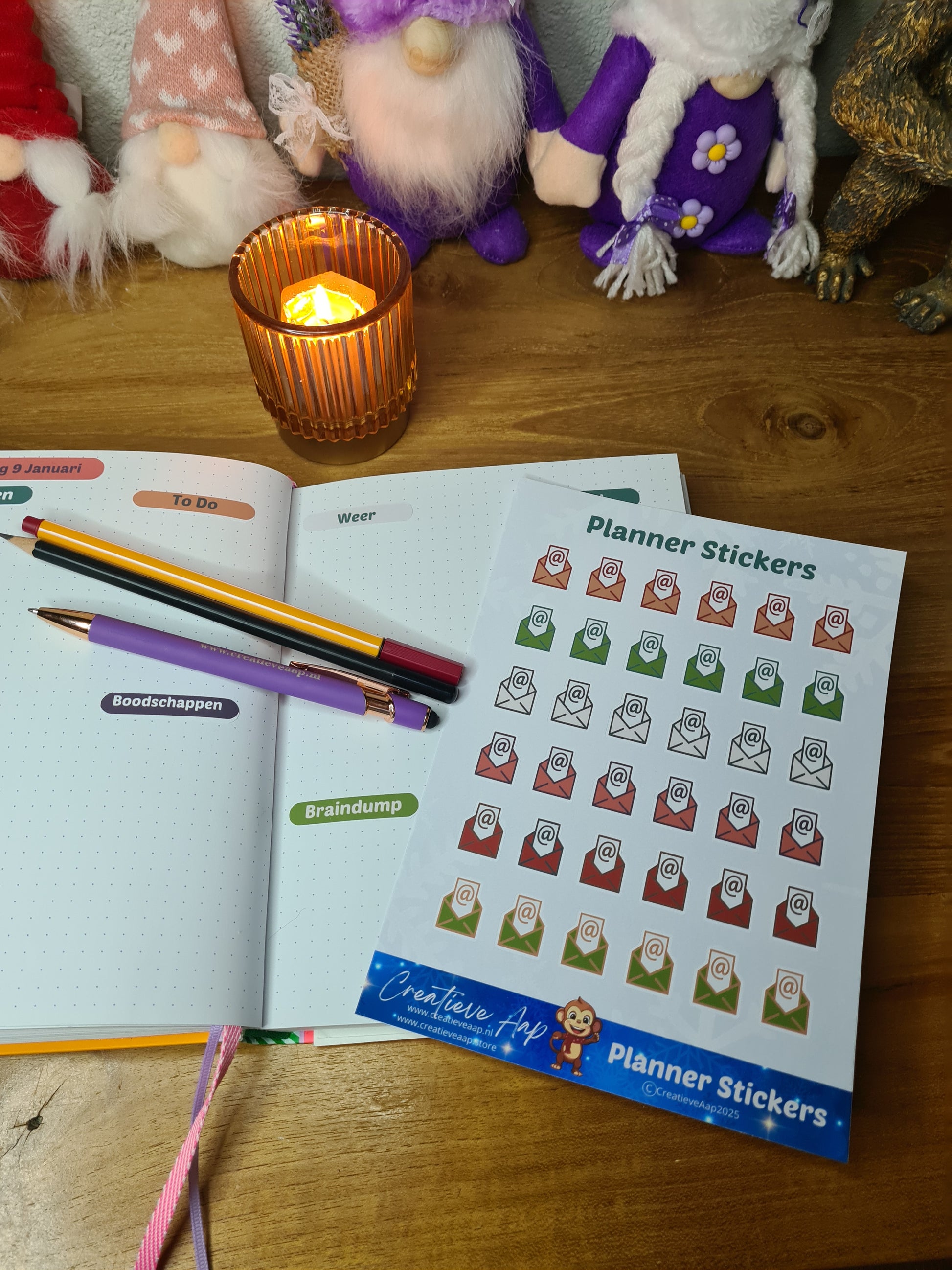 Handgemaakte planner stickers met email icoon in diverse kleuren, beschikbaar in A6 en A5, met keuze uit matte of glanzende afwerking. Ideaal voor het plannen en organiseren van e-mails en digitale taken in planners of bullet journals. Te zien in een warme en gezellige setting met kaarslicht en creatieve accessoires.