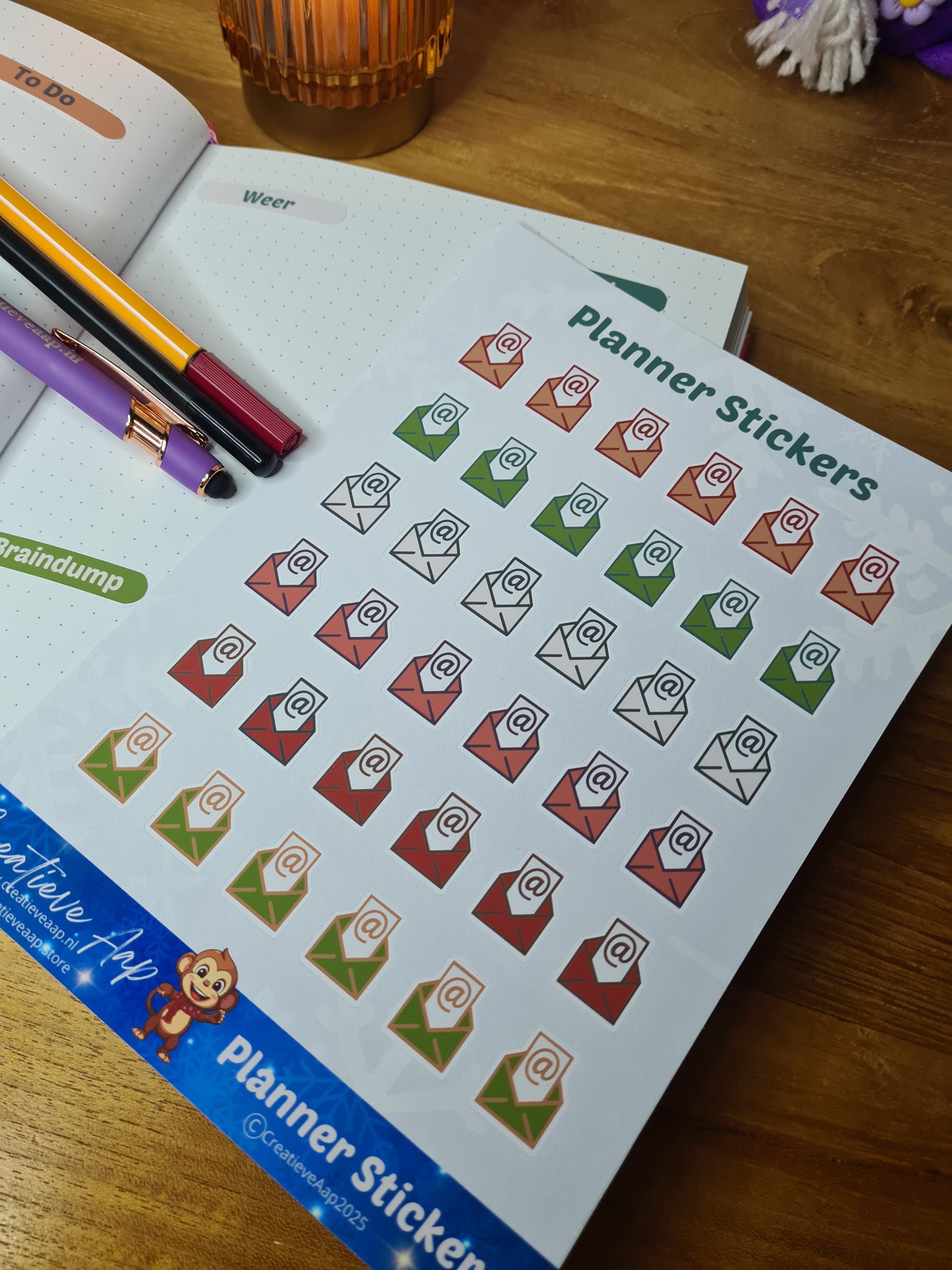 Handgemaakte planner stickers met email icoon in diverse kleuren, beschikbaar in A6 en A5, met keuze uit matte of glanzende afwerking. Ideaal voor het plannen en organiseren van e-mails en digitale taken in planners of bullet journals. Te zien in een warme en gezellige setting met kaarslicht en creatieve accessoires.