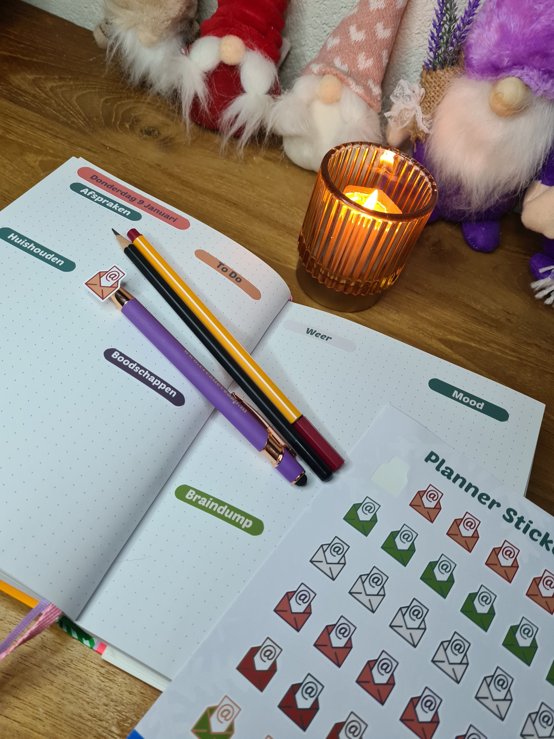 Handgemaakte planner stickers met email icoon in diverse kleuren, beschikbaar in A6 en A5, met keuze uit matte of glanzende afwerking. Ideaal voor het plannen en organiseren van e-mails en digitale taken in planners of bullet journals. Te zien in een warme en gezellige setting met kaarslicht en creatieve accessoires.