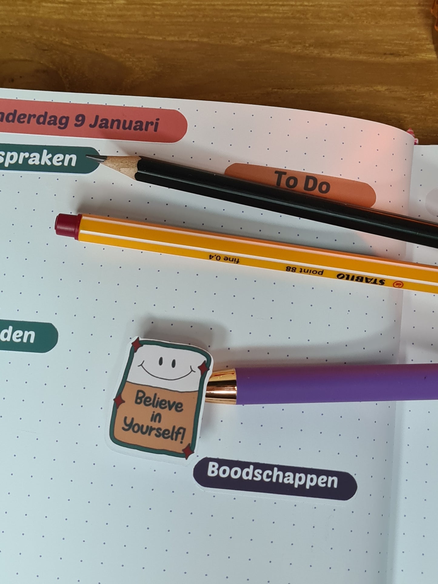 Close-up van een sticker met de tekst 'Believe in Yourself' in een open planner, naast kleurrijke pennen en secties voor planning.