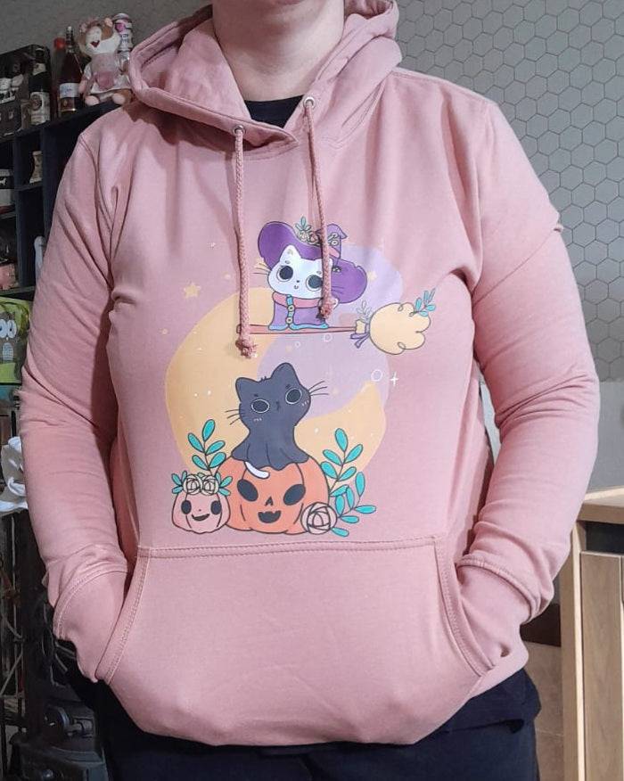 "Persoon draagt een roze hoodie met een schattige Halloween-afbeelding van een zwarte kat die op een pompoen zit, omringd door bladeren en kleine pompoenen. Boven de kat vliegt een cartoon skeletfiguur verkleed als heks op een bezemsteel voor een gele maan."