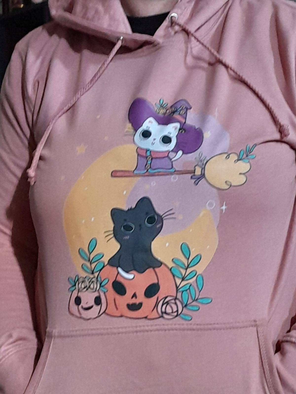 "Persoon draagt een roze hoodie met een schattige Halloween-afbeelding van een zwarte kat die op een pompoen zit, omringd door bladeren en kleine pompoenen. Boven de kat vliegt een cartoon skeletfiguur verkleed als heks op een bezemsteel voor een gele maan."