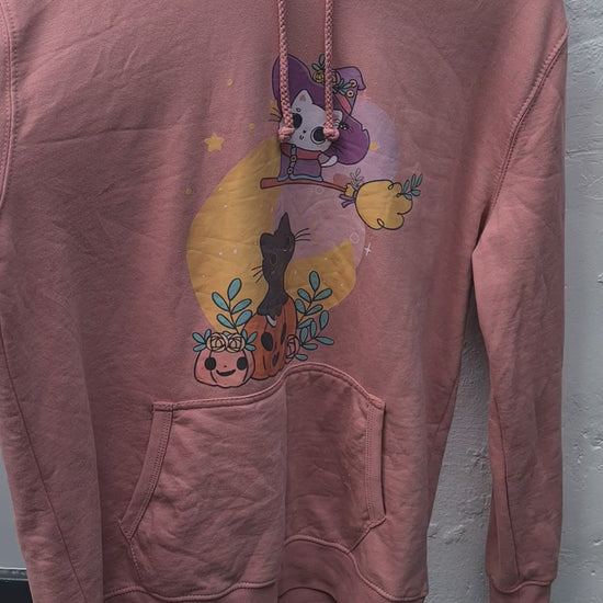 "Persoon draagt een roze hoodie met een schattige Halloween-afbeelding van een zwarte kat die op een pompoen zit, omringd door bladeren en kleine pompoenen. Boven de kat vliegt een cartoon skeletfiguur verkleed als heks op een bezemsteel voor een gele maan."