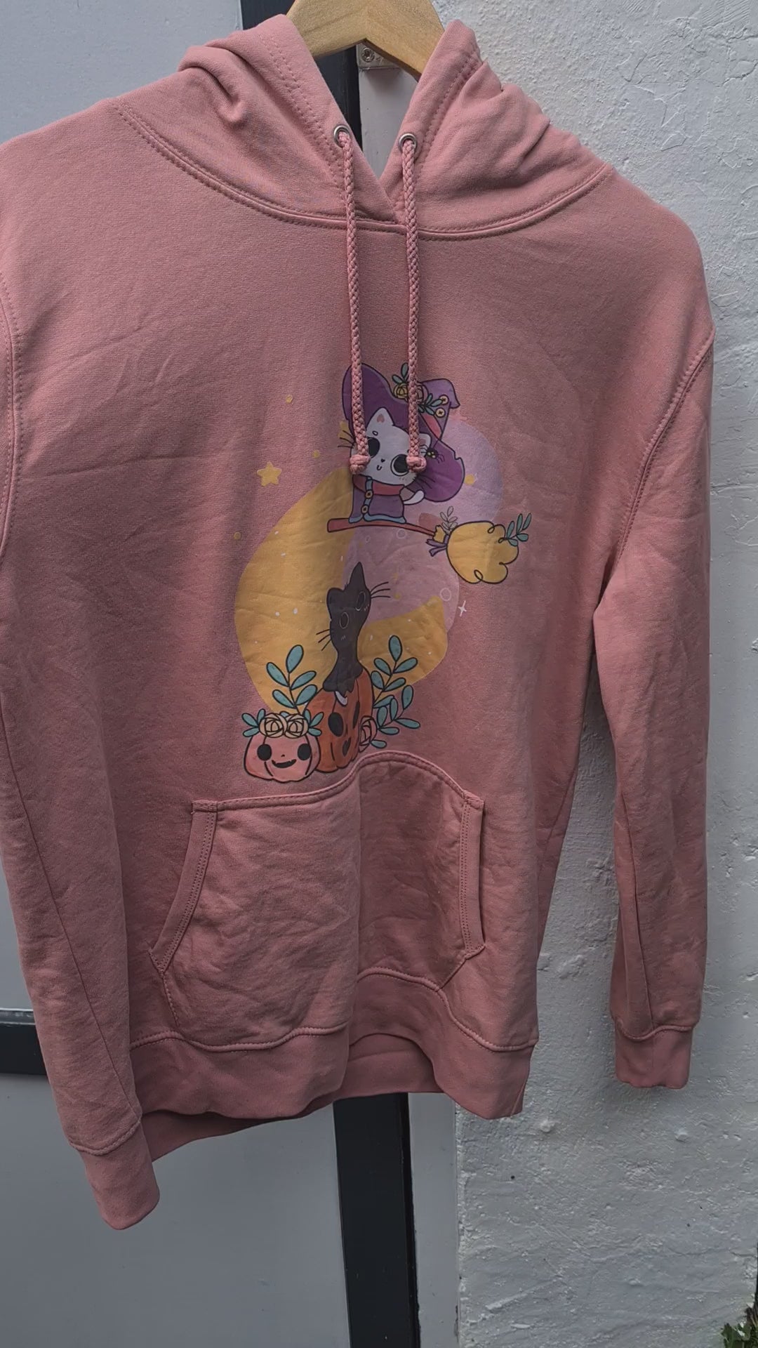 "Persoon draagt een roze hoodie met een schattige Halloween-afbeelding van een zwarte kat die op een pompoen zit, omringd door bladeren en kleine pompoenen. Boven de kat vliegt een cartoon skeletfiguur verkleed als heks op een bezemsteel voor een gele maan."